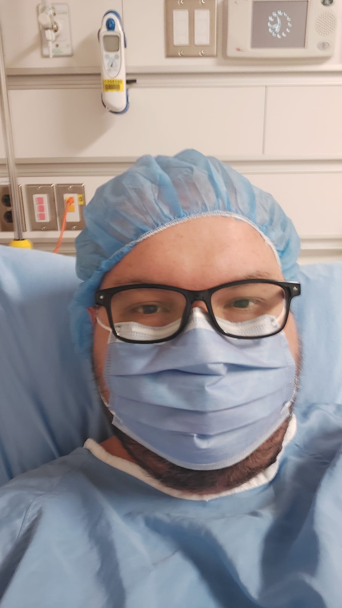 A man is in a hospital bed, wearing a hospital gown, surgical mask and hair covering, he looks nervous